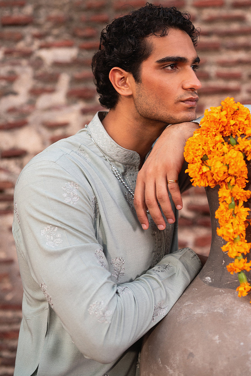 Man in traditional attire leaning against a brick wall with marigold flowers.