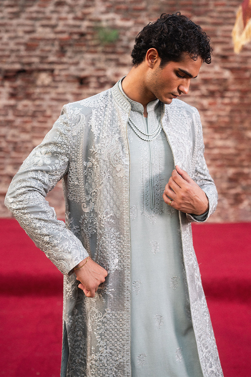 Man wearing a silver embroidered sherwani against a brick wall.